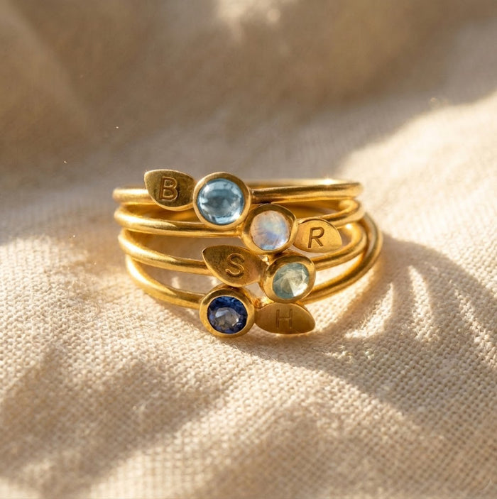 Stackable birthstone initial rings in gold with 3mm faceted gemstones and stamped leaf charms, personalized for family, mom, or couples.
