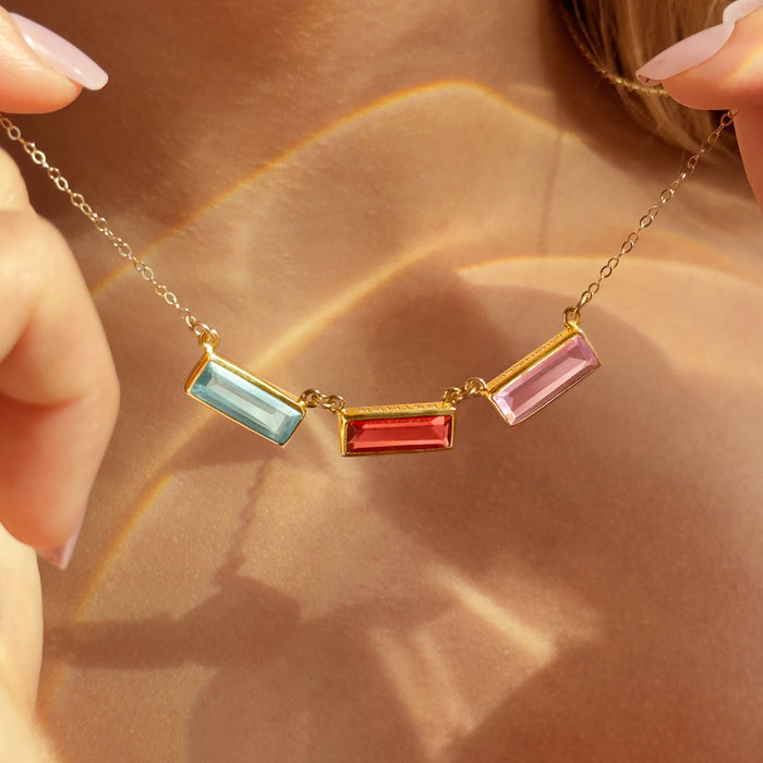 Adira baguette birthstone bar necklace for mom featuring three rectangular bezel-set gemstones on a delicate gold chain. Personalized engraving available with names, initials, dates, or a hidden message. Shown layered for everyday wear, this custom birthstone mom necklace is a meaningful gift for Mother’s Day, birthdays, or Christmas. Stones pictured include aquamarine, ruby, and rose hues; also available in sterling silver and rose gold options for grandma or new moms.