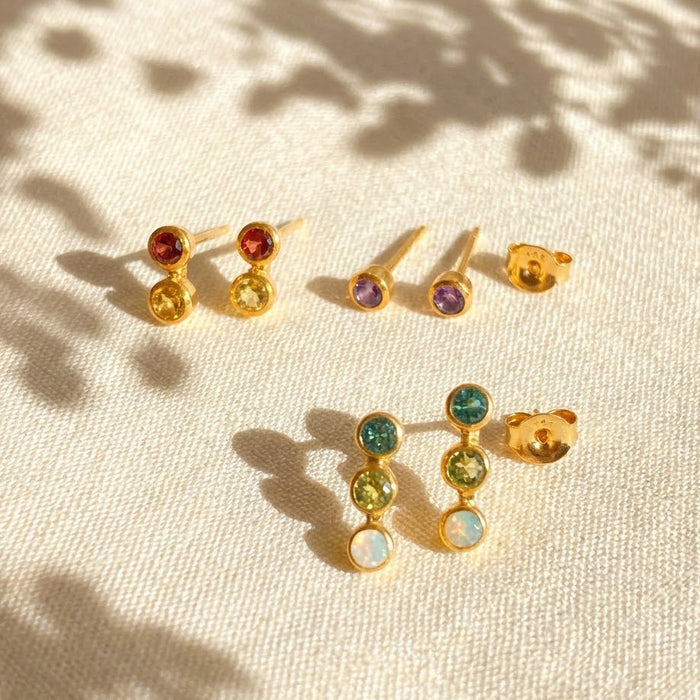 Custom birthstone bar stud earrings with one, two, and three gemstone options in 18K yellow gold, shown on soft fabric with natural shadows for a dainty personalized jewelry look.