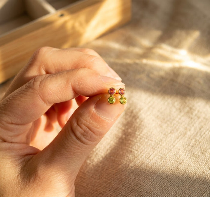 Close-up of hand holding colorful custom birthstone bar studs, showing tiny personalized gemstone earrings made in sterling silver or 18K gold.