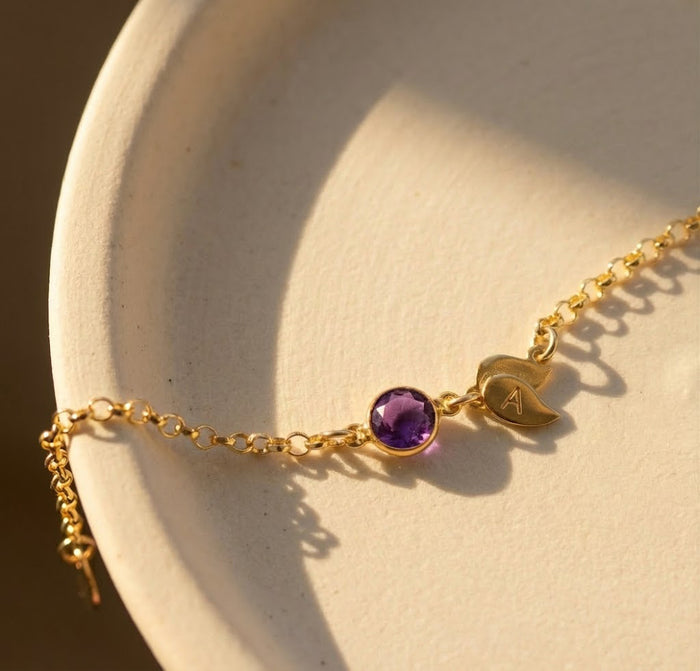 Close-up of personalized birthstone bracelet with pink gemstone and engraved initial leaf charm, adjustable gold bracelet with dainty chain and lobster clasp