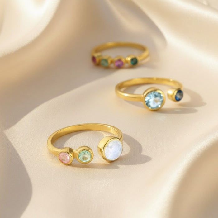 Custom birthstone adjustable rings for moms displayed on soft champagne satin, featuring smooth gold open bands with bezel-set gemstones. Front ring shows a three-stone family design with one larger glowing moonstone for mom and two smaller pastel birthstones for children, while the background rings showcase other colorful multi-gem options. Meaningful personalized family jewelry, perfect Mother’s Day, birthday or Christmas gift for moms who love dainty gold rings.
