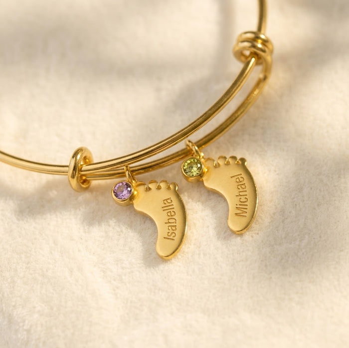 Gold custom mom bangle bracelet with engraved footprint charms, children’s names, and tiny faceted birthstones on a warm neutral background.