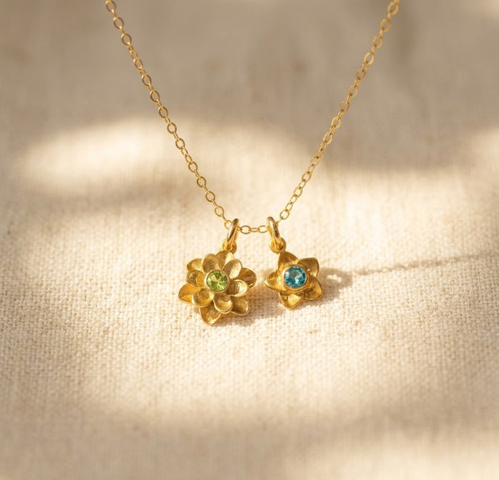 Gold birthstone flower charm necklace on warm beige fabric with colorful gemstone centers, meaningful personalized necklace for mom.