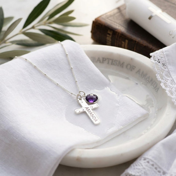 Personalized cross necklace with engraved pendant and purple birthstone charm styled on white dish