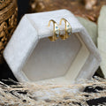 Gold hoop earrings with blue topaz accents on a white hexagonal stand with decorative elements.