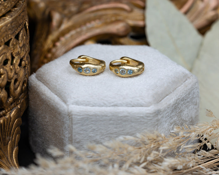 Gold hoop earrings with blue topaz accents on a white hexagonal stand with decorative elements.