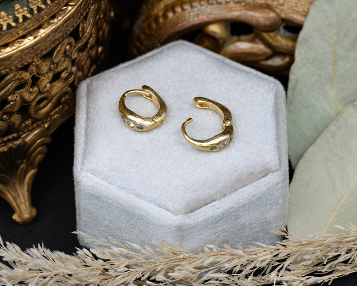 Gold hoop earrings with blue topaz accents on a white hexagonal stand with decorative elements.