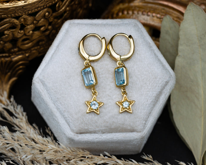 Gold earrings with star and blue topaz gemstone accents on a decorative cushion.