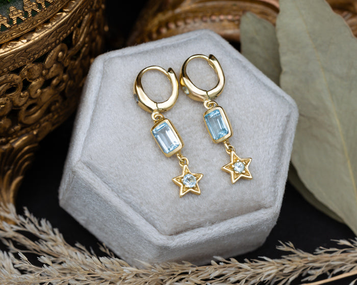 Gold earrings with star and blue topaz gemstone accents on a decorative cushion.