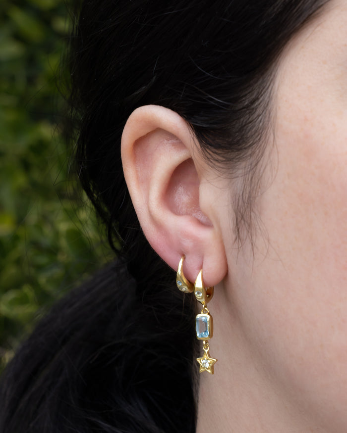 Close-up of an ear wearing gold earrings with blue topaz accents against a blurred green background