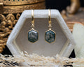 Gold earrings with hexagon moss agate on a decorative stand.