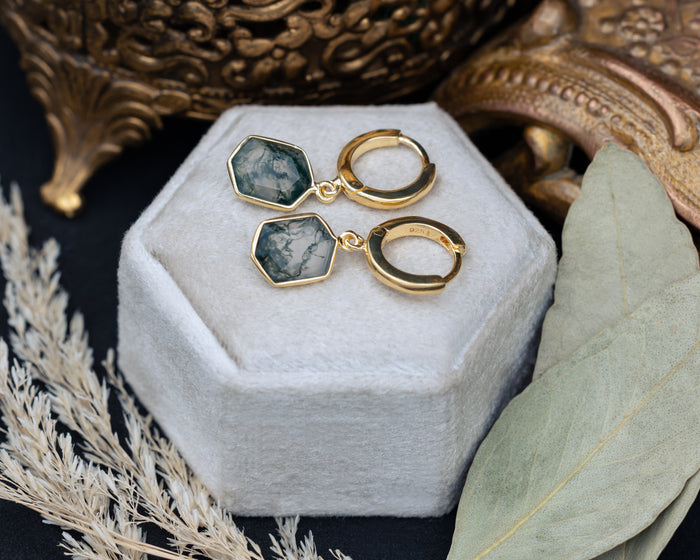 Gold earrings with moss agate on a white jewelry box with decorative elements.