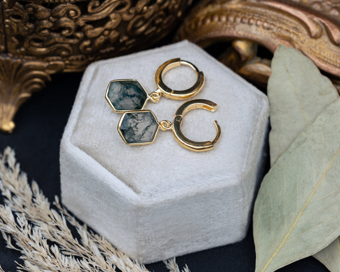 Gold earrings with moss agate on a white jewelry box with decorative elements.
