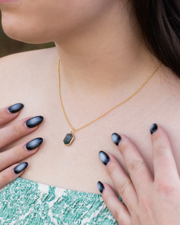 Gold necklace with a moss agate pendant worn by a person with dark nail polish.
