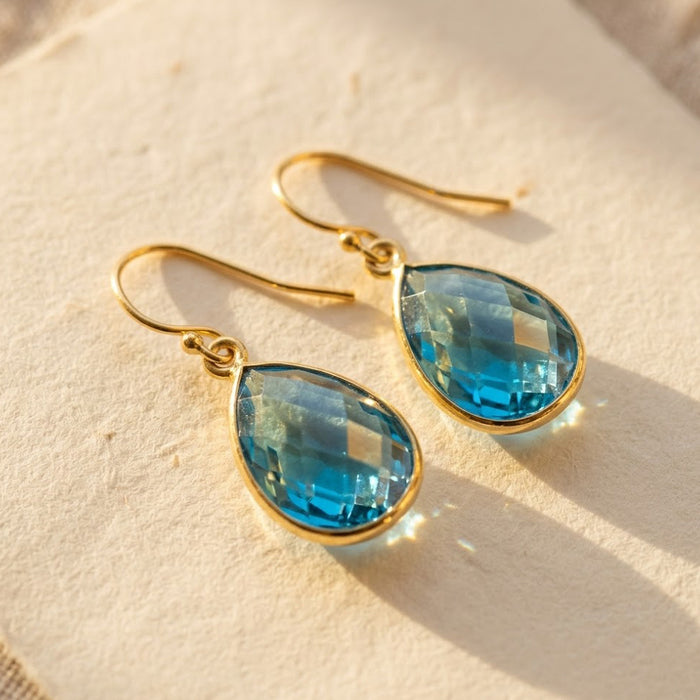 London blue topaz teardrop earrings in 14k gold filled on beige background, faceted December birthstone drop earrings