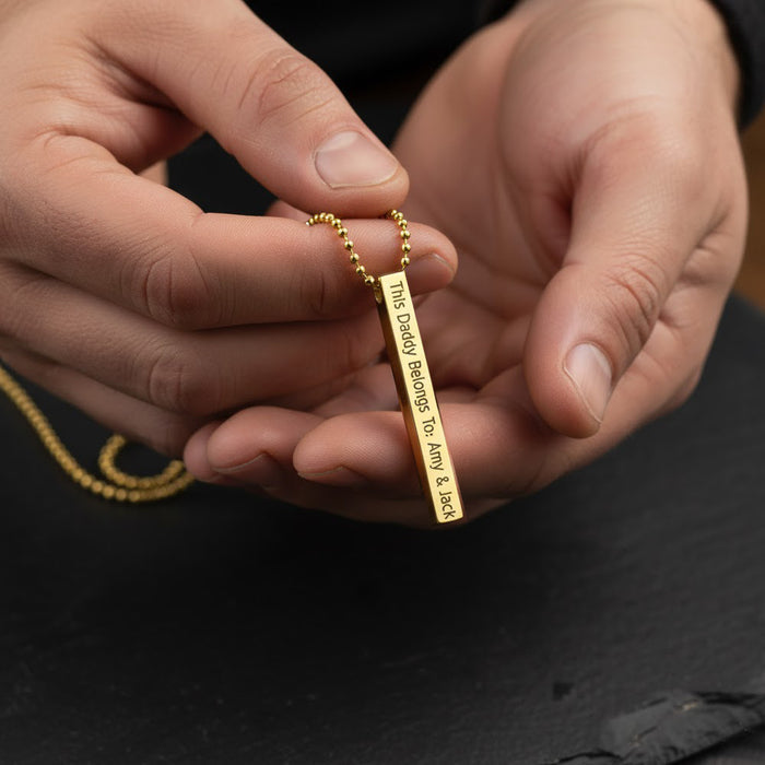 Gold custom engraved 3D bar necklace for men held in both hands, featuring a vertical bar pendant on a beaded chain with personalized message “This Daddy Belongs To…” and children’s names. Modern masculine jewelry that can be customized with names, dates or coordinates, perfect Father’s Day, birthday or new dad gift from kids or partner.