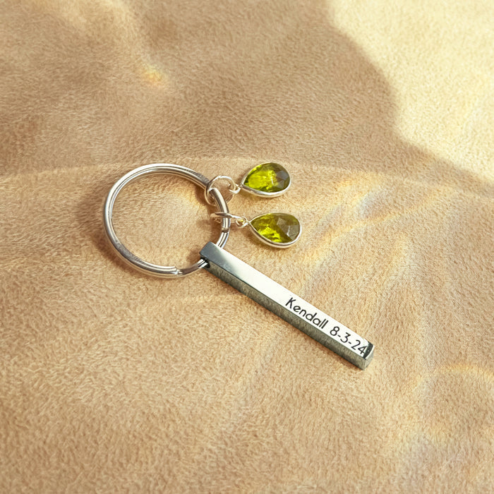 Close view of a silver engraved mom keychain resting on warm fabric, featuring a slim 3D bar etched with a short message and accented by two teardrop birthstone charms. The petite size keeps pockets light while allowing up to four sides of personalization—names, dates, roman numerals, or GPS coordinates. A thoughtful mother’s day keychain and everyday keepsake for moms of two.