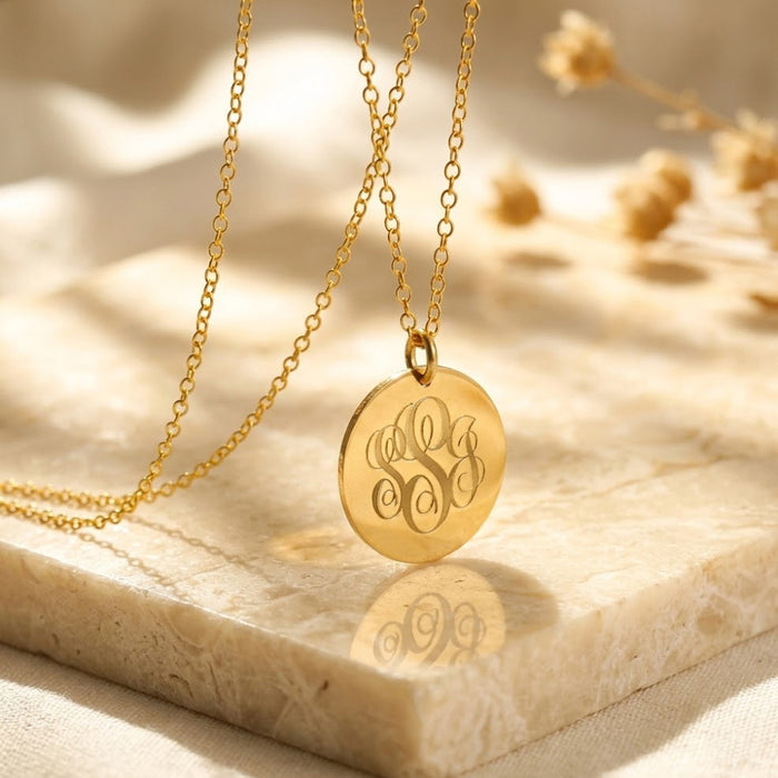 Close-up of a gold monogram disc necklace on soft fabric, featuring engraved initials on a round pendant with delicate chain.