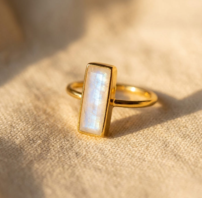 Natural moonstone bar ring in gold with a rectangular June birthstone gemstone, shown as a dainty minimalist ring on soft fabric.