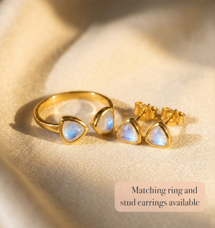 Gold moonstone triangle ring with matching stud earrings shown on warm fabric.