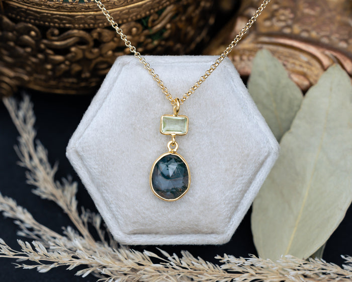 Gold necklace with moss agate & prasiolite pendant on a hexagonal white velvet stand with decorative elements.