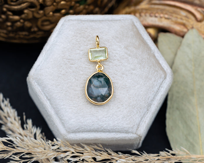 Gold pendant with green gemstones on a hexagonal white velvet stand.