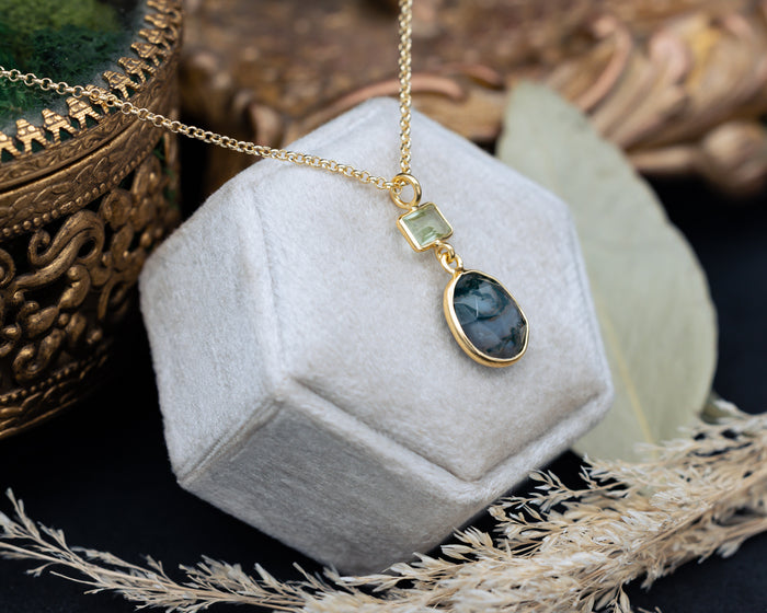 Gold necklace with moss agate & prasiolite pendant on a hexagonal white velvet stand with decorative elements.