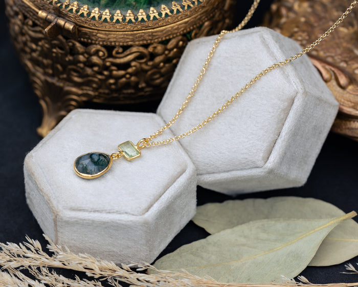 Gold necklace with moss agate & prasiolite pendant on a hexagonal white velvet stand with decorative elements.