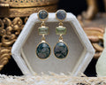 Gold earrings with green gemstones on a white stand with a decorative background