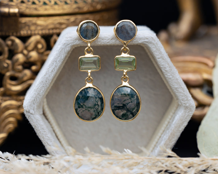 Gold earrings with green gemstones on a white stand with a decorative background