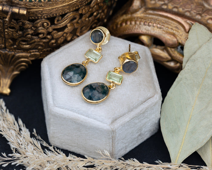 Gold earrings with green gemstones on a white jewelry box with decorative elements.