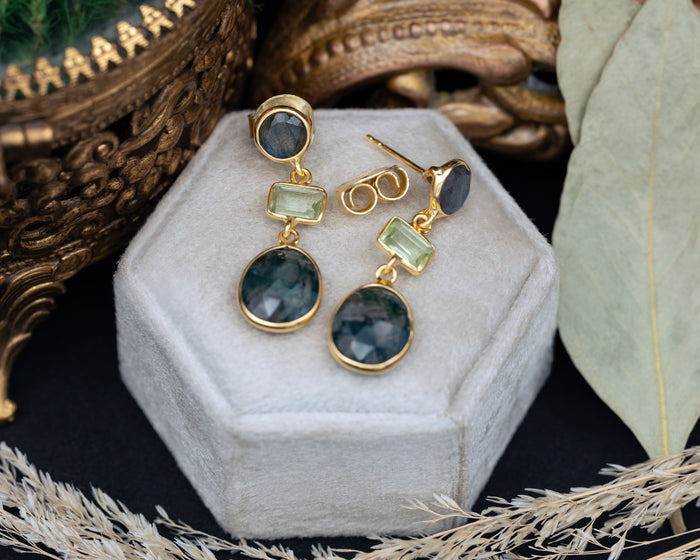 Gold earrings with green gemstones on a white hexagonal stand with decorative background