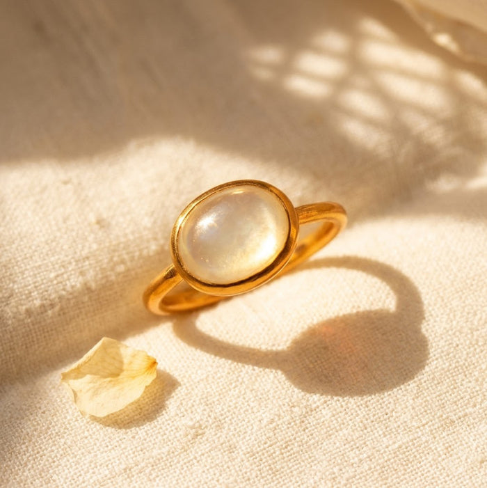 Oval gemstone ring shown on beige background with mother of pearl cabochon center stone and simple minimalist band.