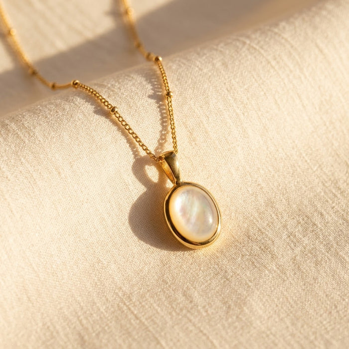 Mother of Pearl oval pendant necklace in gold shown on soft cream fabric in natural sunlight.