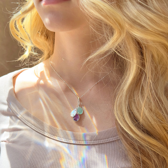 Dainty custom birthstone mom necklace with prong-set teardrop stones shown on a blonde woman in natural sunlight, emphasizing the sparkle and rich color of each birthstone. Elegant layered look that pairs with casual outfits or special occasions, personalized with children’s or family birth months, a thoughtful gift for mom, grandma, or best friend who loves meaningful jewelry.