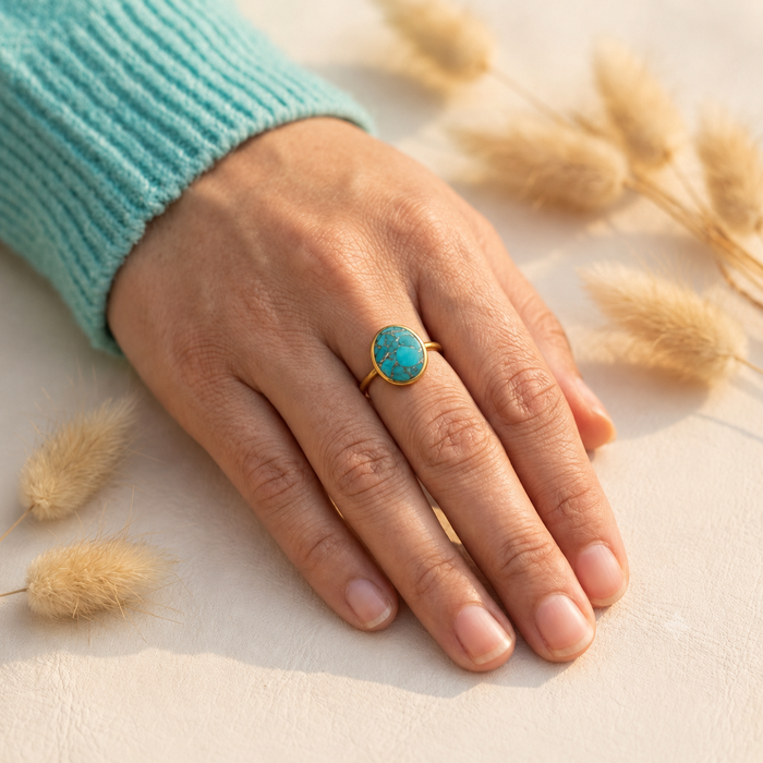 Woman’s hand wearing a gold oval turquoise ring beside dried grasses on a softly lit neutral surface.