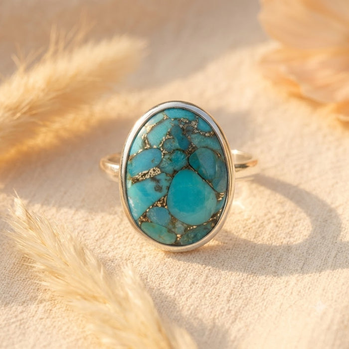 Close-up of an oval turquoise ring with natural copper matrix displayed on cream fabric with dried botanical accents.
