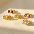 Oval birthstone stud earrings shown on beige fabric with garnet, green amethyst, purple amethyst, aqua chalcedony, and lemon quartz gemstones.
