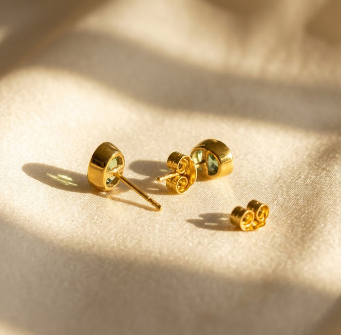 Pair of oval birthstone stud earrings shown on beige fabric with matching earring backs in warm natural light.