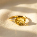 Lemon quartz oval ring shown on warm beige fabric with a pale yellow 8 x 10mm gemstone in a gold bezel setting.