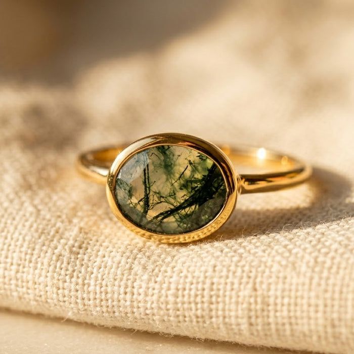 Oval moss agate ring in gold vermeil displayed on soft beige fabric, showing the faceted green gemstone and delicate stacking band.