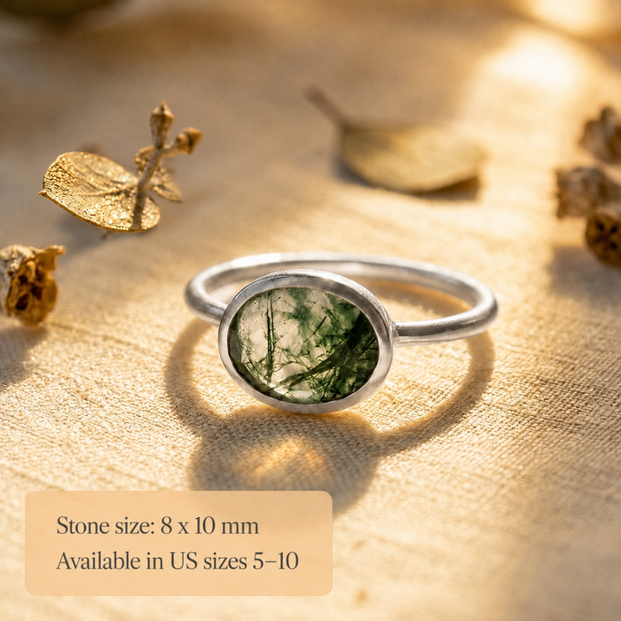 Sterling silver moss agate ring with oval 8x10mm green gemstone shown on warm textured background with soft shadow.