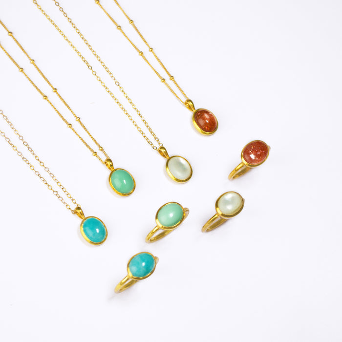 Four Gold Necklaces with oval cabochon pendants with matching Gold rings below. From Left to Right: Blue Amazonite on a cable chain, mint green Chrysoprase on a satellite chain, Mother of Pearl on a cable chain, and Sunstone on a satellite chain