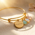 Gold personalized birthstone bangle bracelet with engraved mama disc charm and three faceted birthstones on a warm neutral background.