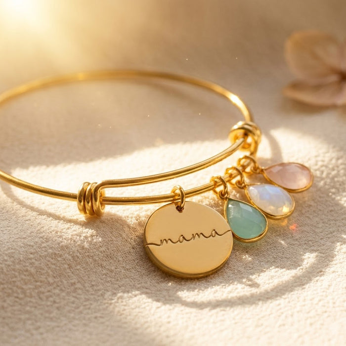 Gold personalized birthstone bangle bracelet with engraved mama disc charm and three faceted birthstones on a warm neutral background.