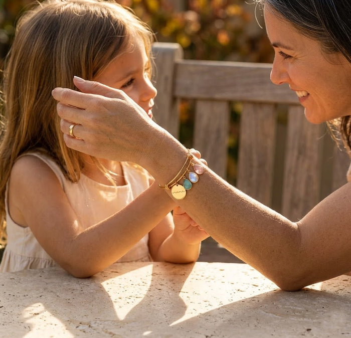 Custom family birthstone bangle bracelet worn by mom hugging her daughter, featuring engraved name disc charms and small faceted birthstones.