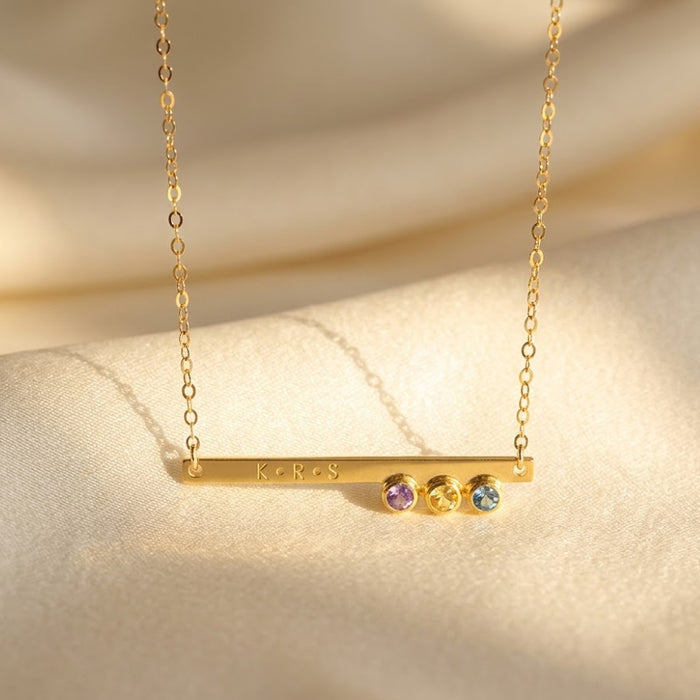 Dainty gold birthstone bar necklace with engraved name and tiny gemstone charms on beige fabric