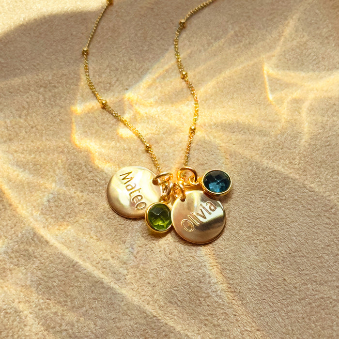 Personalized gold disc name necklace with two engraved charms reading “Mateo” and “Olivia” and matching blue and green birthstone crystals on a dainty beaded chain, custom family jewelry gift for mom or grandma, sentimental mothers necklace keepsake.