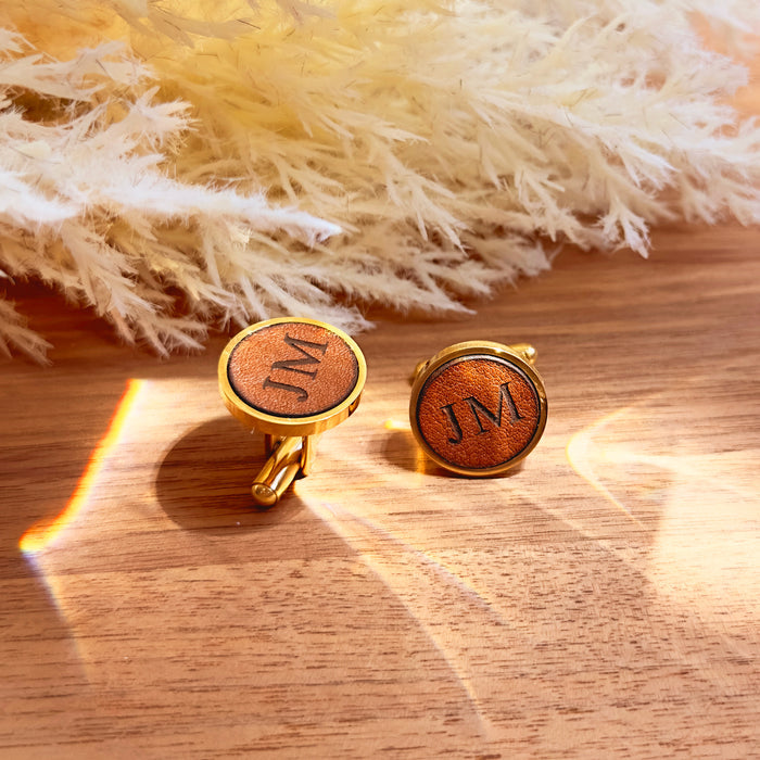 Custom engraved leather cufflinks with JM initials, round brown leather in gold bezel on wood surface with warm sunlight, personalized monogram cufflinks for groom, husband or groomsmen, unique wedding, anniversary or Father’s Day gift for him.